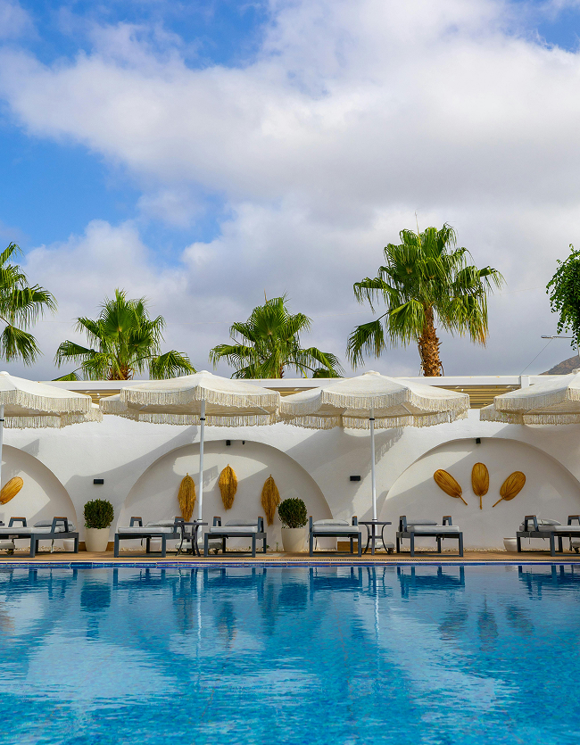 Resort swimming pool lined with lounge chairs, fringed umbrellas, and palm trees against a white arched wall